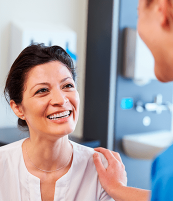 Paciente mulher sorrindo sendo acolhida por profissional de medicina. Programa de apoio ao paciente crônico gerencia a saúde de pacientes com determinadas doenças crônicas e os auxilia nos tratamentos em geral e em eventuais complicações. Fornece orientações, esclarecimentos e suporte emocional por telefone.