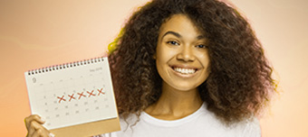 Moça de cabelos cacheados e bastante sorridente mostrando um calendário marcado em sua mão.