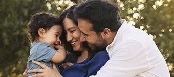 Família feliz composta por pai, mãe e filho pequeno, sorrindo e abraçados em um momento de carinho ao ar livre.